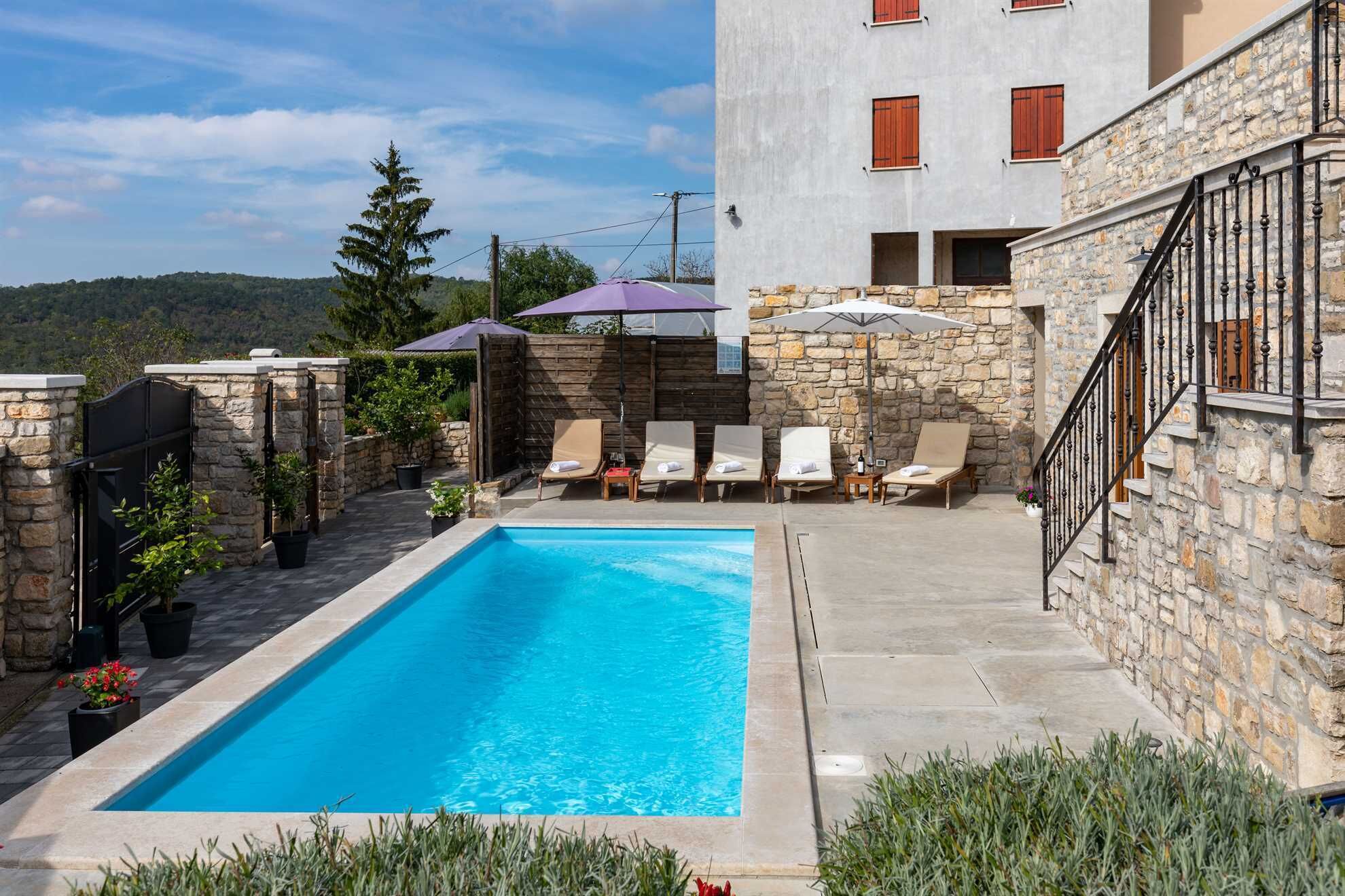 Una piscina con sillones de descanso y sombrillas, rodeada de muros de piedra y un edificio con contraventanas rojas.