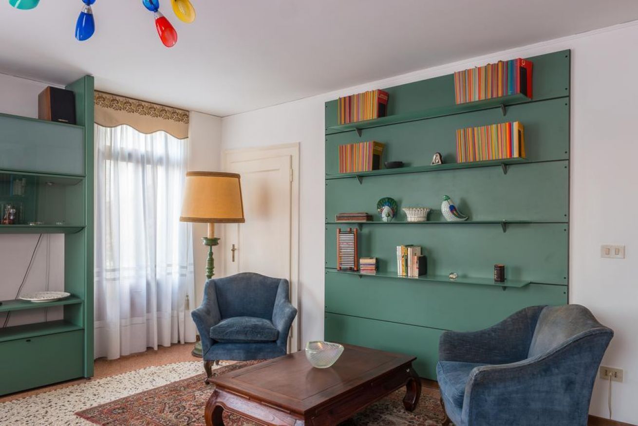 Ein Wohnzimmer mit einem grünen Bücherregal, zwei blauen Sesseln, einem Holztisch und einer Lampe.