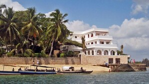 A white multi-story building with a beachside location, palm trees, and boats on the water. - Luxury beachfront holiday house, pool garden all sea views, Forodhani House Lamu (Lamu)