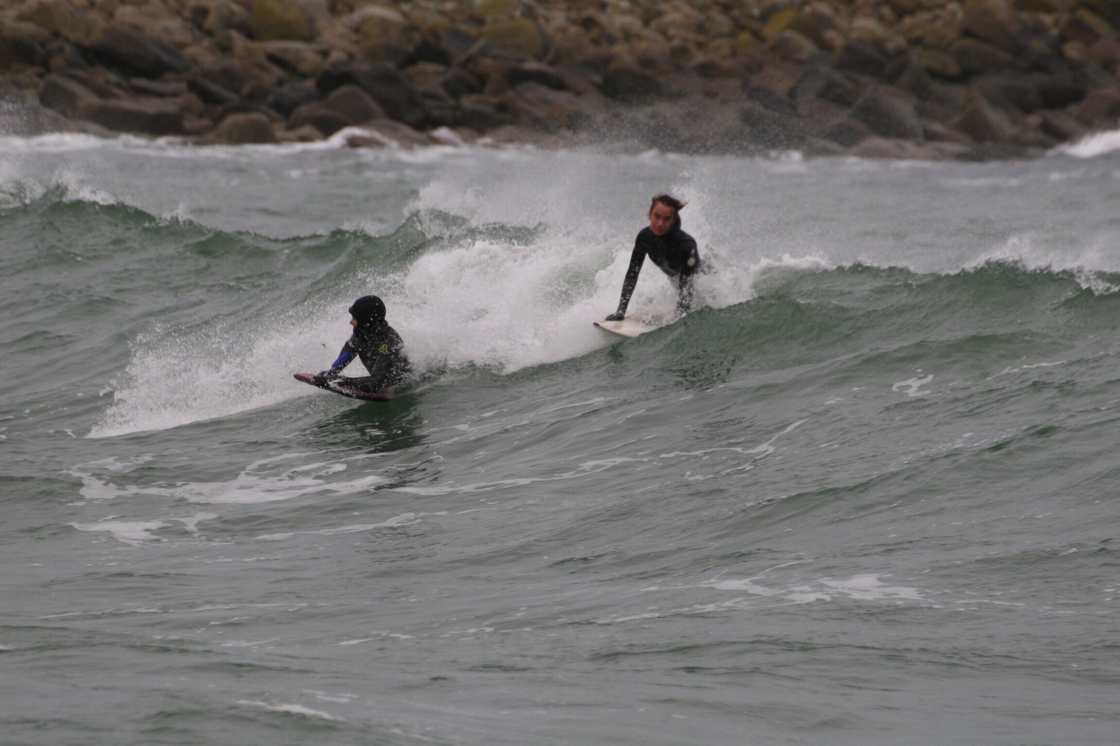 Two surfers on a wave with a rocky shoreline in the background.