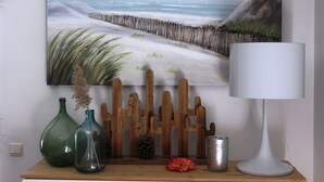 Una pintura de una escena de playa con una cerca de madera, dos jarrones verdes, una flor roja y una lámpara blanca sobre un estante de madera.