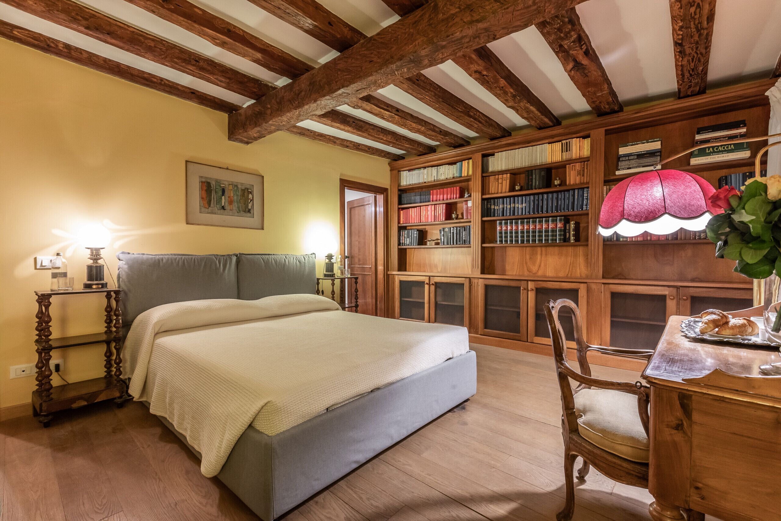 A bedroom with a bed, wooden bookshelves, a desk with a lamp, and a wooden chair.