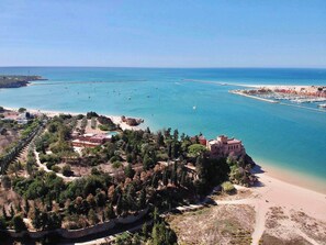 Uma área costeira com praia, um porto de iates e um edifício histórico rodeado de árvores.