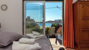 Een slaapkamer met een bed, een houten kledingkast, een balkon met uitzicht op zee en gebouwen, en een glazen schuifdeur.