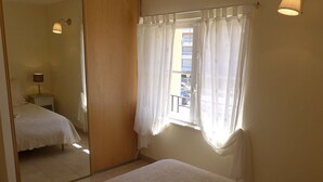 Ein Schlafzimmer mit einem Bett, einem Nachttisch mit Lampe, einem Fenster mit Vorhängen und einem Spiegel, der den Raum reflektiert.