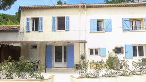 Ein zweistöckiges Gebäude mit blauen Fensterläden, einer überdachten Eingangspforte und einem Garten mit Pflanzen.