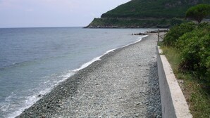 Een kiezelstrand met een betonnen pad ernaast, dat leidt naar een groene heuvel met een klif.