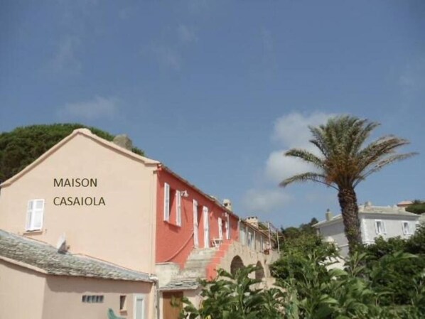 En byggnad med skylten ”MAISON CASAIOLA”, en röd fasad och vita fönster, omgiven av grönska och palmer.