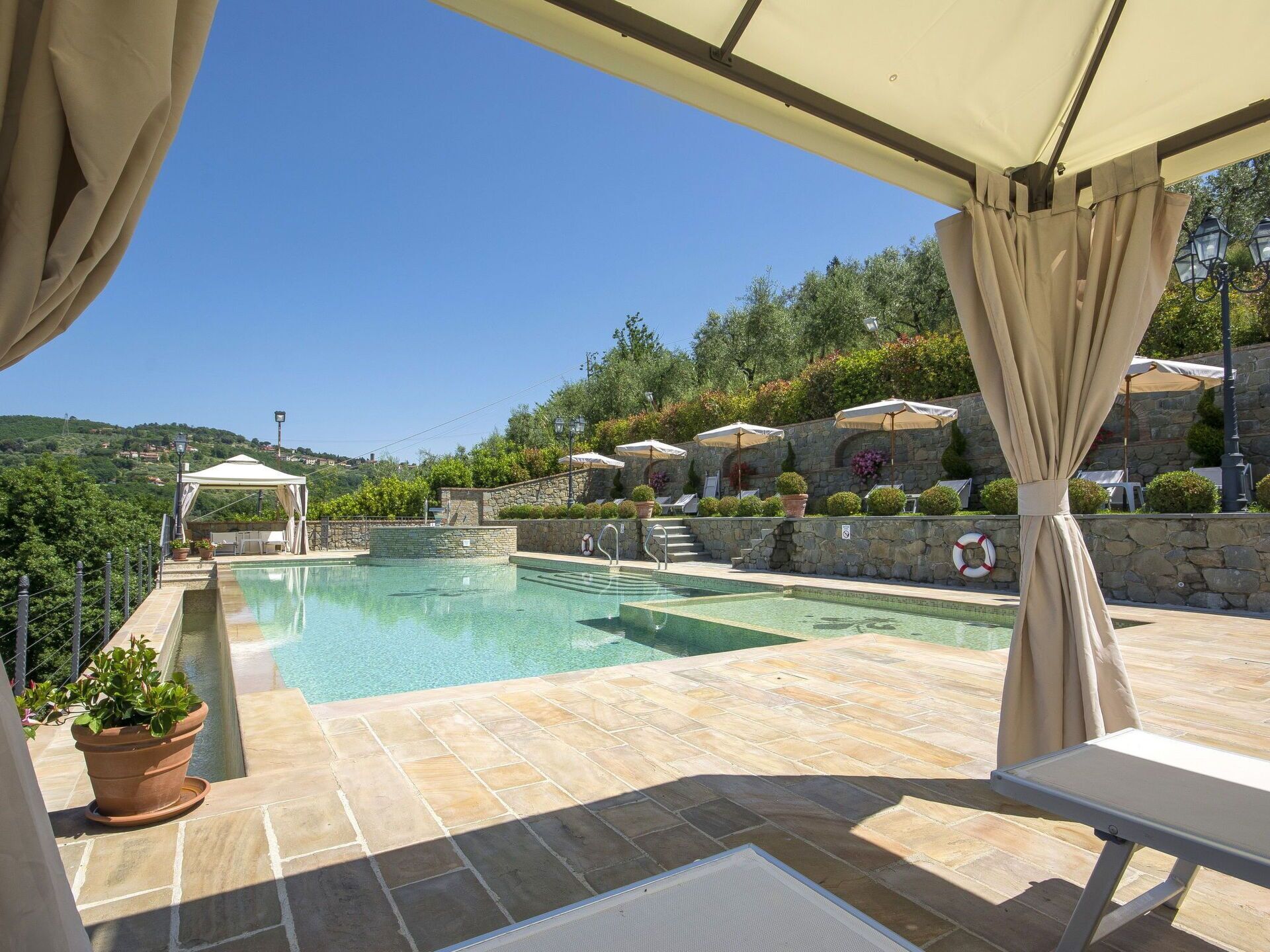 A clear blue swimming pool with a stone deck, surrounded by a stone wall and potted plants. There are lounge chairs and umbrellas under a canopy.