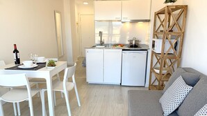 A modern kitchen with white cabinets and a built-in oven, a dining area with white chairs and a table set for two, and a grey sofa with patterned cushions. -  Apartment 1st Line Beach-WIFI-A / A (Empuriabrava)