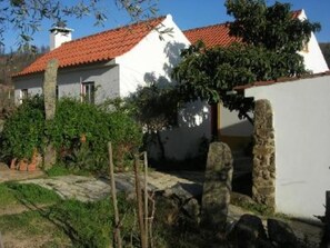Un edificio blanco con techo de tejas rojas, rodeado de follaje y muros de piedra.