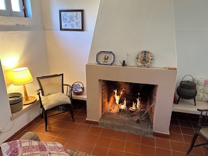 Una sala de estar con chimenea, una silla de mimbre, una mesa con lámpara y un cuadro en la pared.