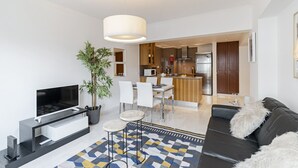A modern living room with a flat-screen TV, a black leather sofa, a dining area with white chairs, and a kitchen in the background. - Arriaga I, amazing location (Funchal)