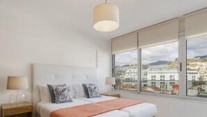 A modern hotel room with a large bed, two bedside tables, a window with a city view, and a hanging light fixture. - Arriaga I, amazing location (Funchal)