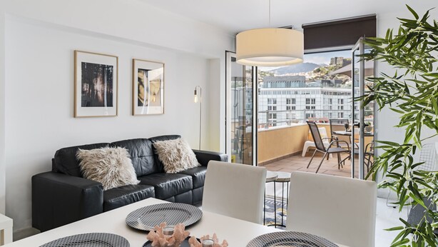 A modern living room with a black leather sofa, a dining table with placemats and decorative items, and a large window offering a city view. - Arriaga I, amazing location (Funchal)