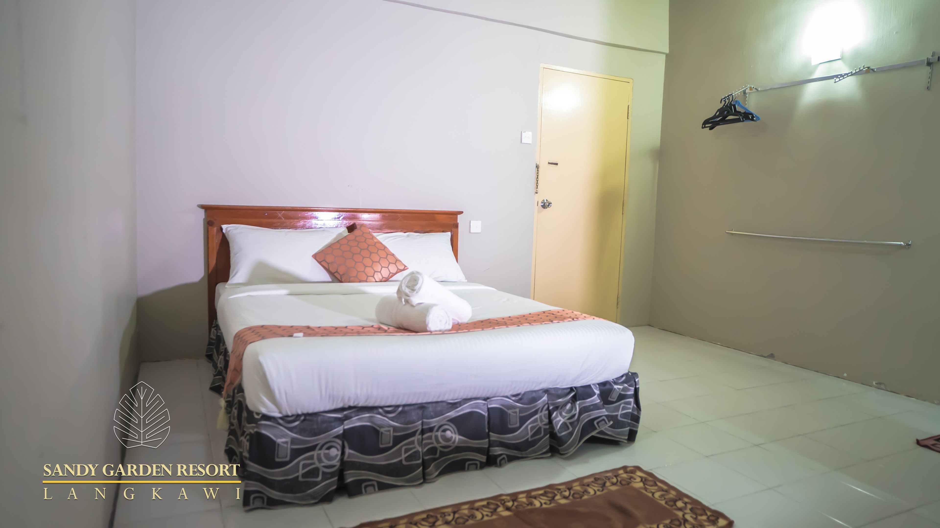 A neatly made bed with a wooden headboard, a patterned bed skirt, and a rolled towel on the foot of the bed. There is a small rug on the floor, a door to the right, and a wall-mounted towel rack above it.