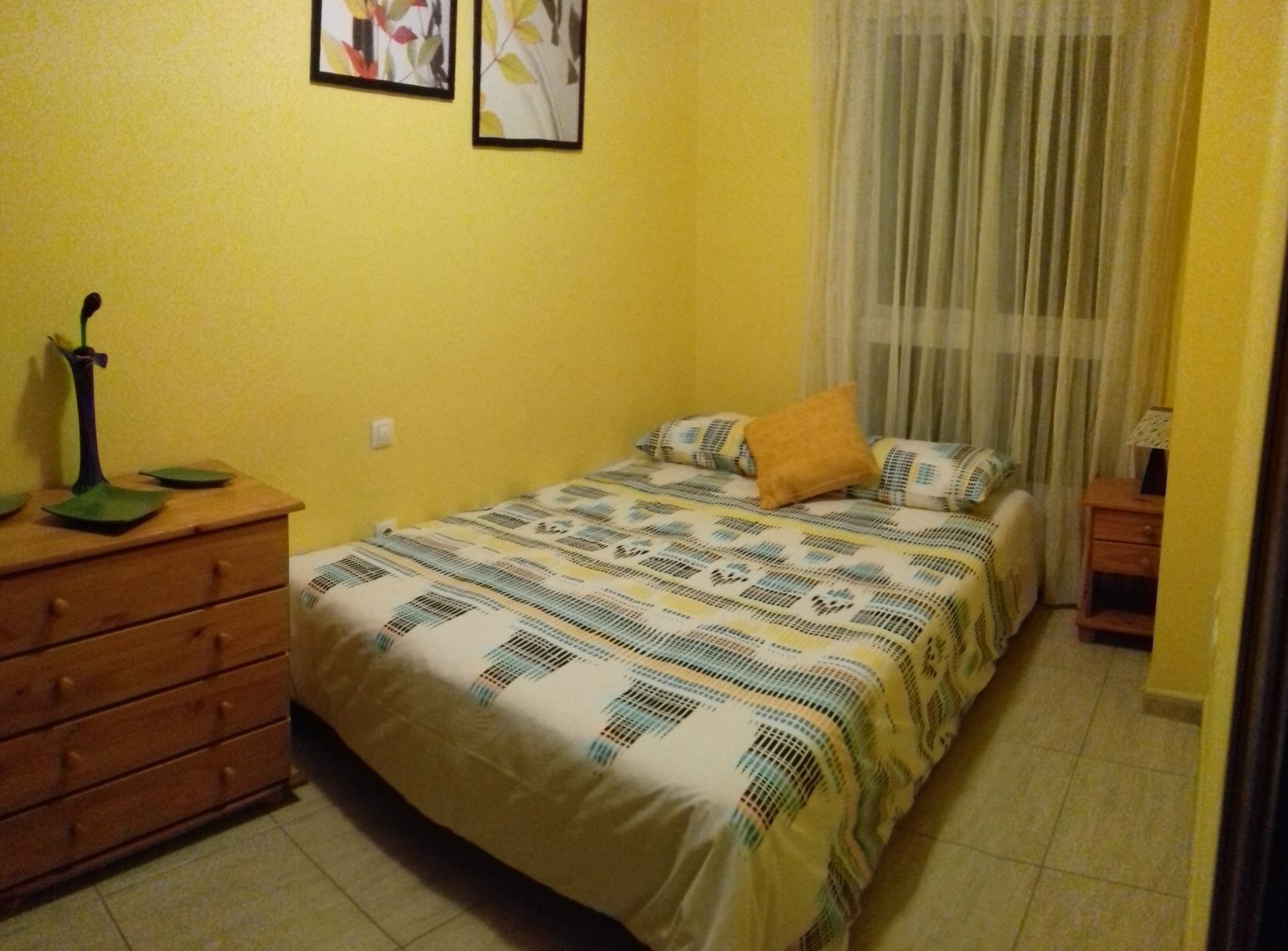 A small bedroom with a bed, a wooden dresser, a nightstand, and a window with curtains.
