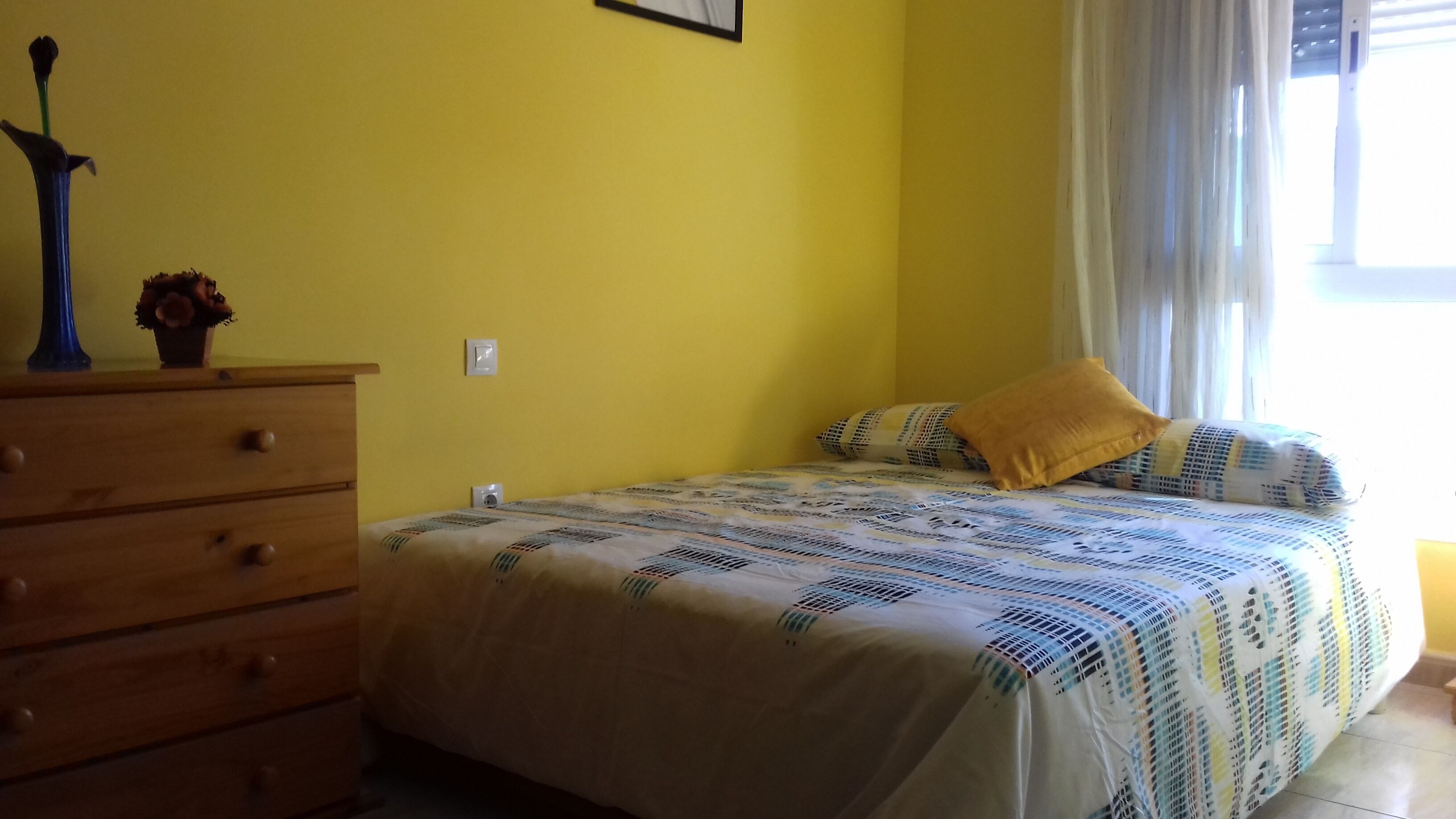 A bedroom with a bed, a dresser, a wall-mounted light switch, and a window with curtains.