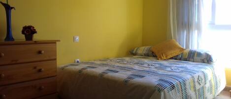 A bedroom with a bed, a dresser, a wall-mounted light switch, and a window with curtains.