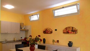 Une cuisine avec une table à manger, des chaises, un vase de fleurs et deux fenêtres fixées au mur.