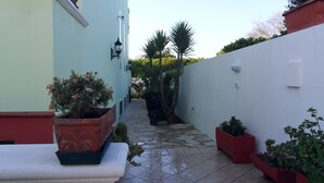 Une petite cour étroite avec un sol carrelé, des plantes en pot et un mur blanc agrémenté d’un luminaire.