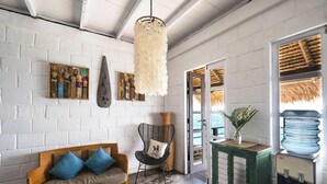 Habitación con pared de ladrillo blanco, sofá de madera con cojines azules, una silla de mimbre, lámpara colgante y dispensador de agua.