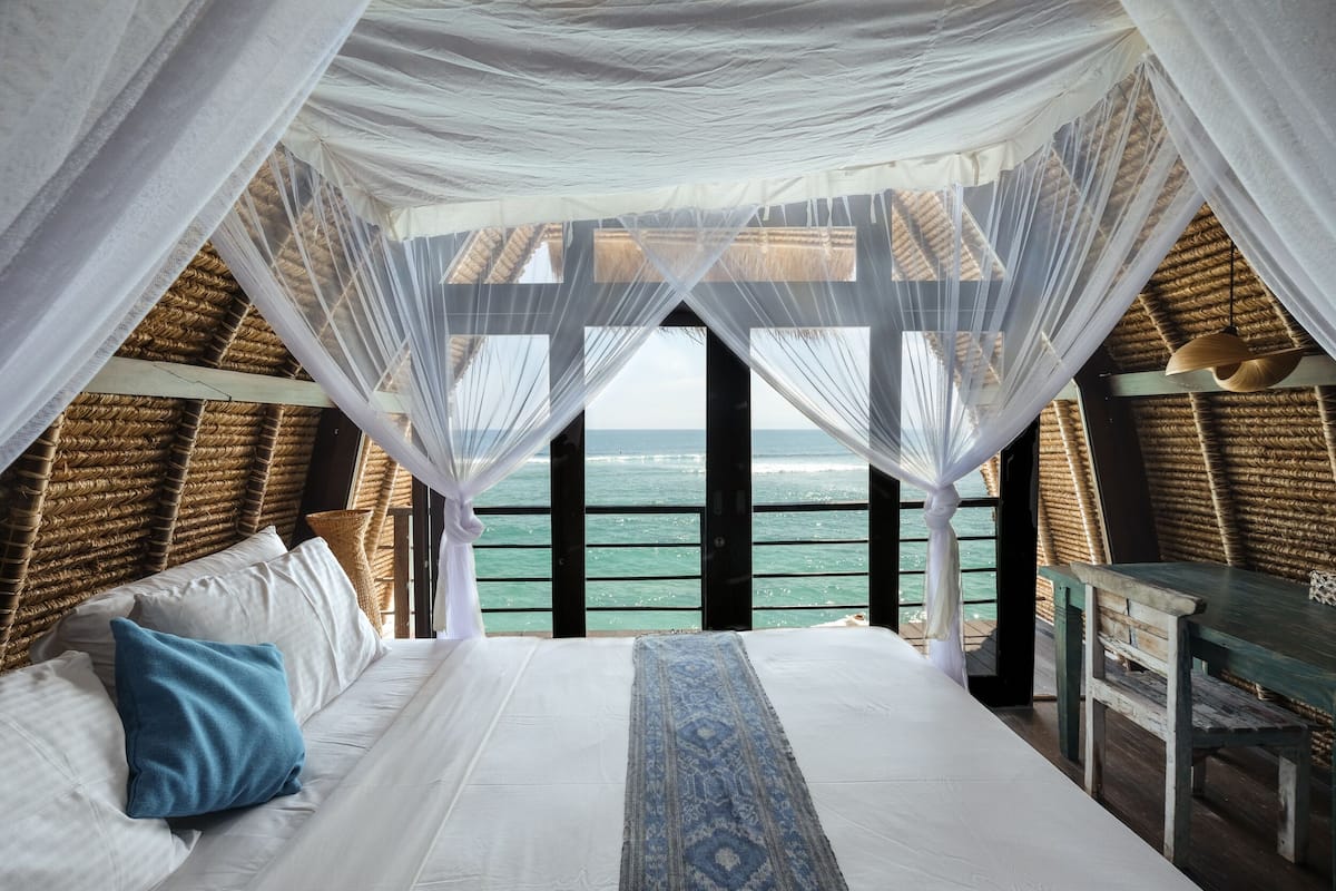 Un dormitorio con una cama grande, ropa de cama blanca, una almohada azul y vista al mar desde un balcón.