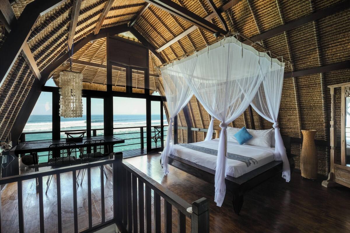 Una habitación con cama bajo un dosel, vista al océano y terraza de madera con mesas y sillas.