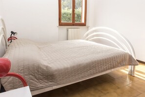A bed with a white headboard, a window with a view of greenery, and a radiator below the window.