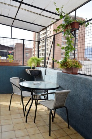 A covered outdoor seating area with a round table and chairs, surrounded by potted plants and a view of buildings. - The Golf with large terrace (Las Condes)