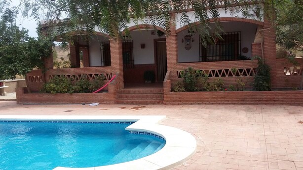 A swimming pool with a brick patio, a covered outdoor seating area, and a brick building with windows and a door. - Holiday home Villa Carmeli (Ardales)