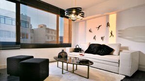 Una sala de estar moderna con un sofá blanco, otomanas negras, una mesa de centro de vidrio y un ventanal con persianas.