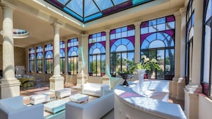 A spacious indoor area with white sofas, a central table, and large windows with stained glass.