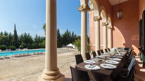 A dining area with a pool view, featuring a long table set for a meal, surrounded by columns and outdoor seating.