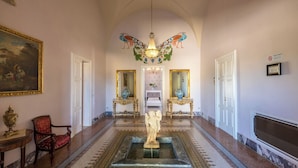 A grand hallway with a marble floor, a statue in the center, and ornate wall decorations including a painting and a chandelier.