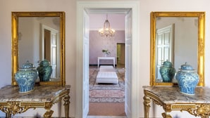 A hallway with two ornate gold-framed mirrors, a white bench, and blue and green vases on marble tables.