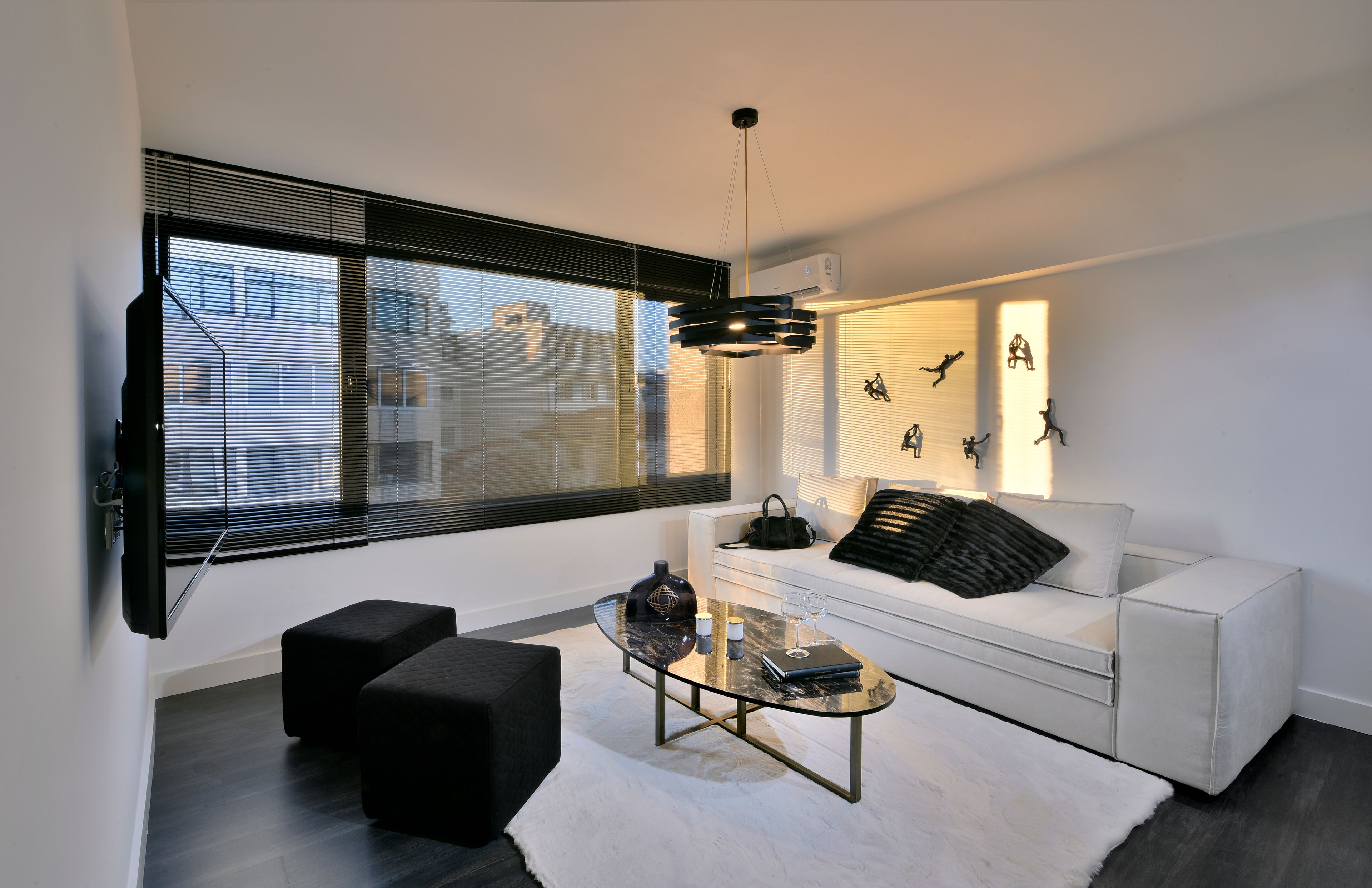 A modern living room with a white sofa, black ottomans, a glass coffee table, and a wall-mounted TV.