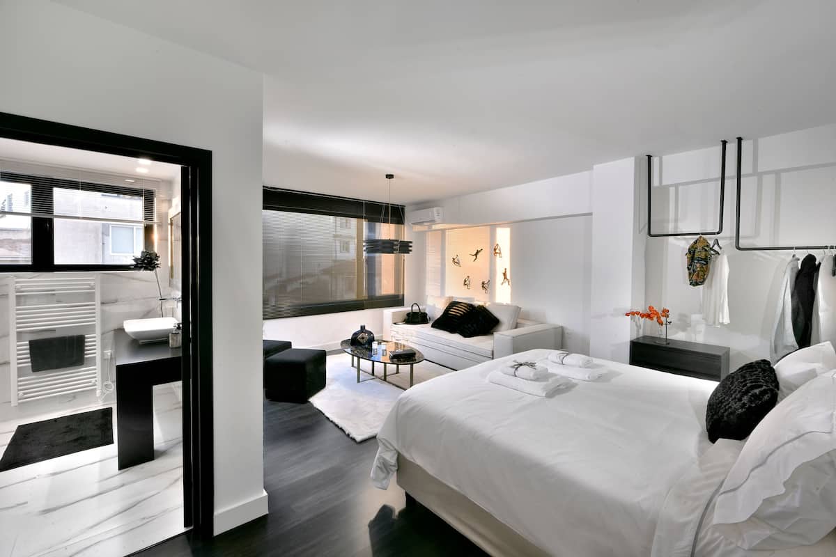 A modern bedroom with a large bed, a sitting area with a sofa and coffee table, and a view of the city through large windows.