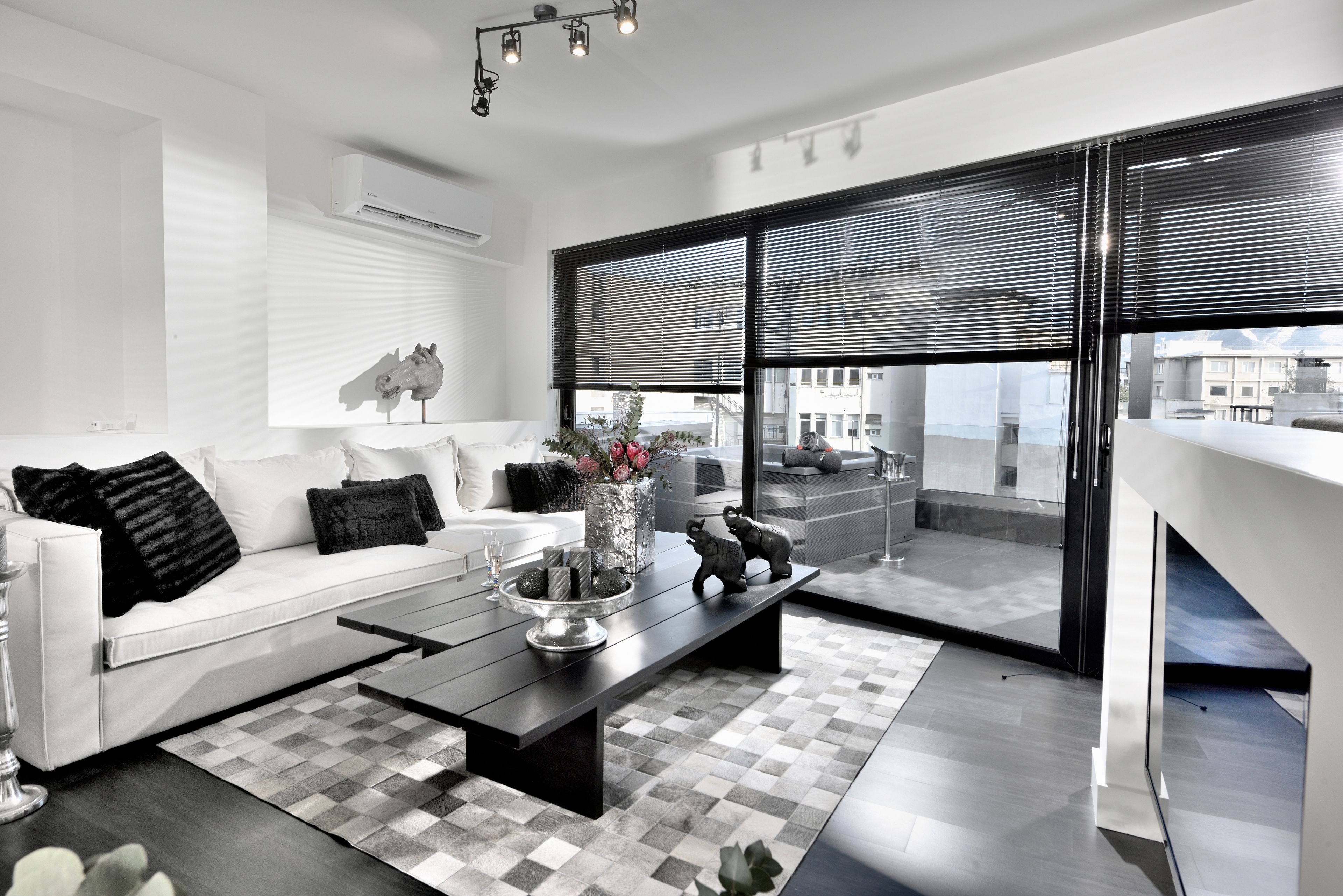 A modern living room with a white sofa, a black coffee table, and a view of the city through large windows with blinds.