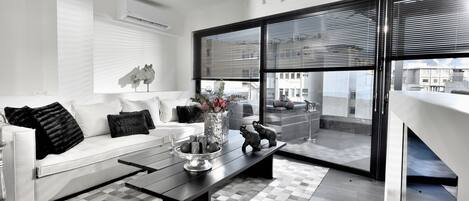 A modern living room with a white sofa, a black coffee table, and a view of the city through large windows with blinds.