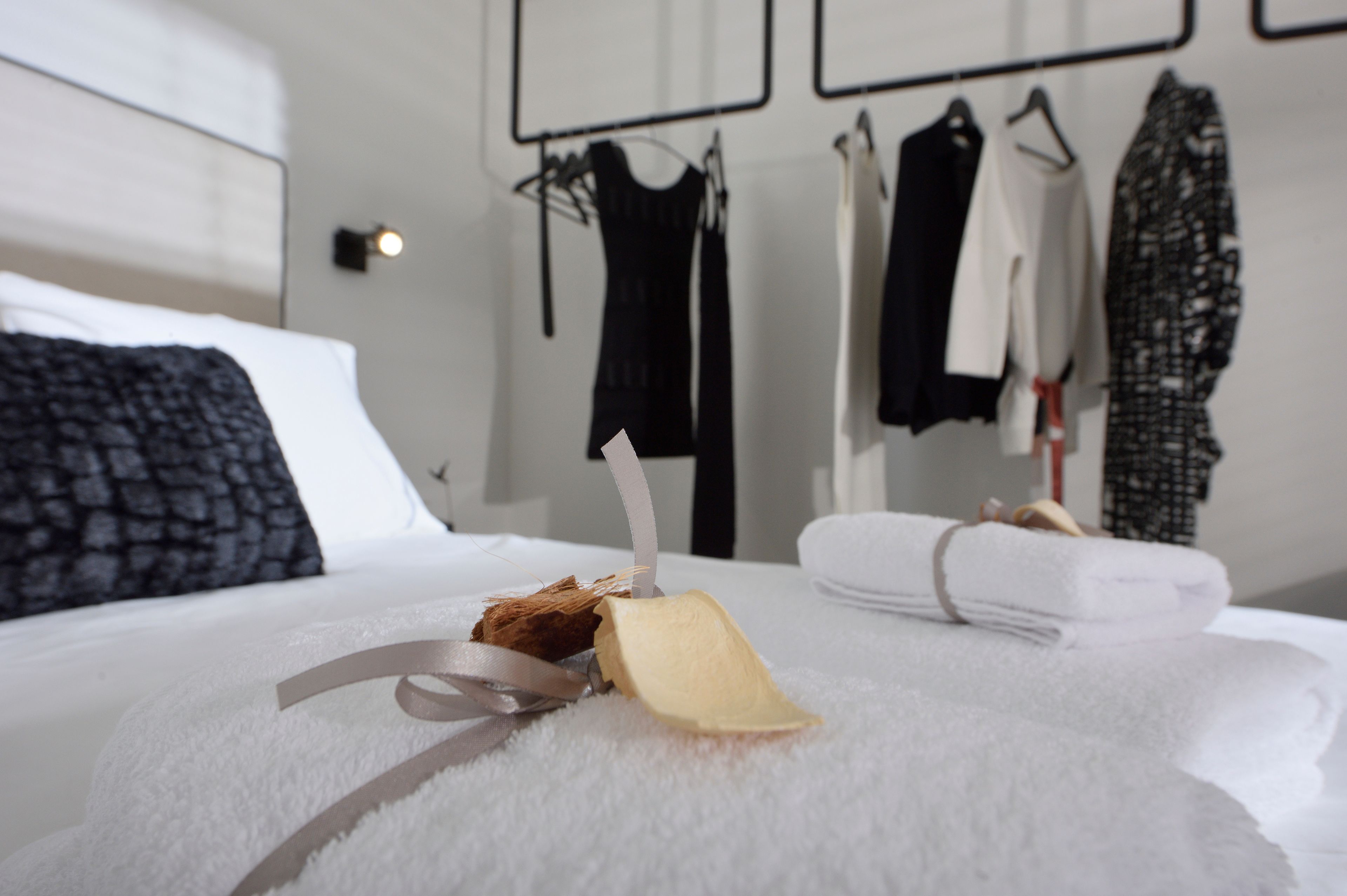 A neatly made bed with a towel and a decorative item on it, a headboard, and a clothing rack with various garments in the background.