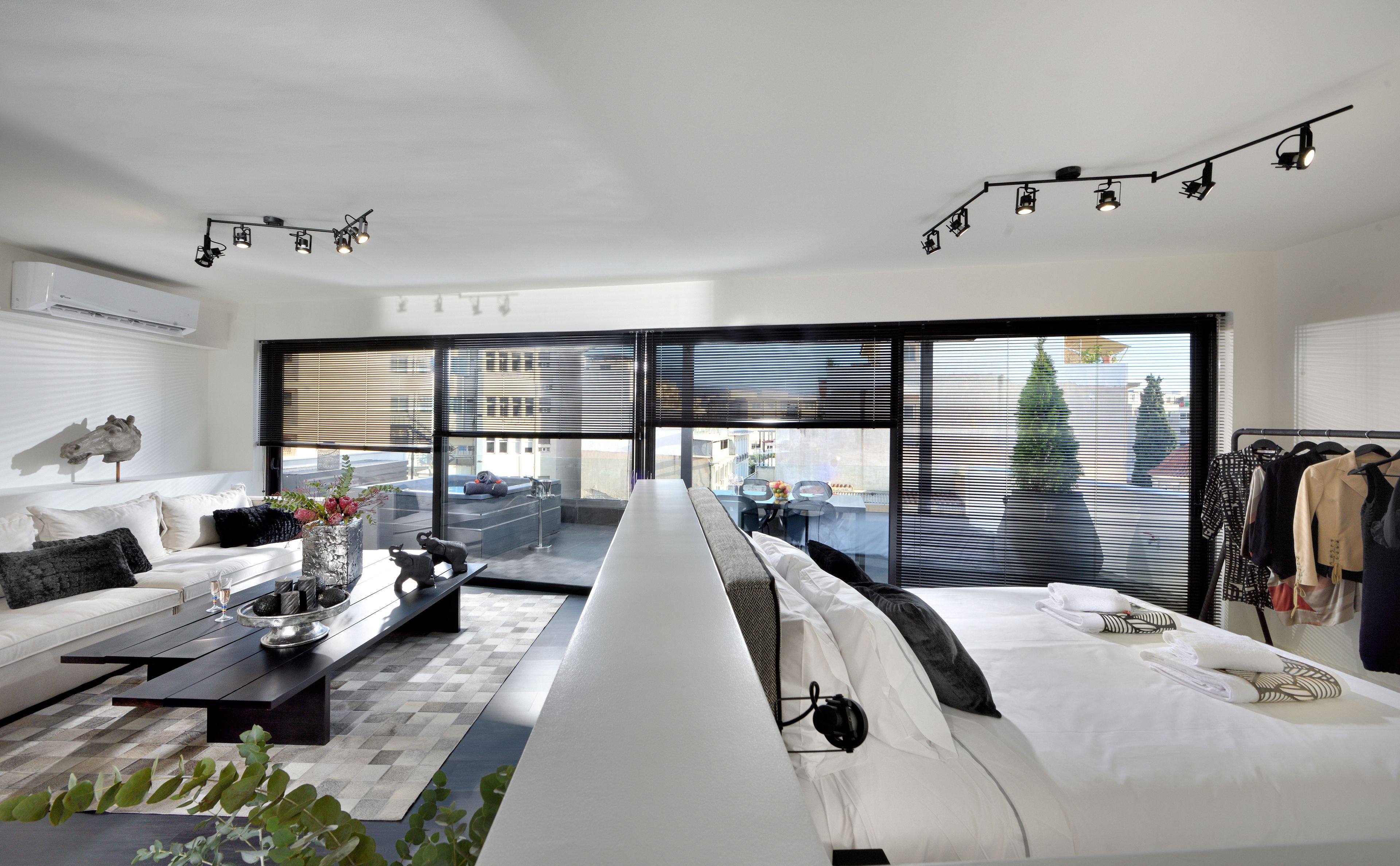 A modern living room with a sofa, coffee table, and a dining area. There is a large window with blinds, a ceiling light fixture, and a view of the city.