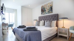 A modern bedroom with a large bed, two bedside tables with lamps, a wall-mounted TV, and a window with curtains. - 1K Waterside - Praia da Luz (Luz)