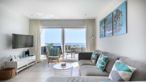 A modern living room with a large flat-screen TV, a grey sofa with teal and white cushions, a wicker basket, and a small round coffee table. - 1K Waterside - Praia da Luz (Luz)