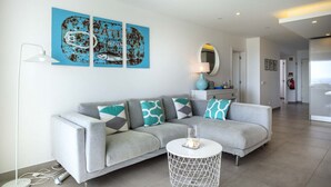 A modern living room with a grey sofa, teal and white patterned cushions, a white side table with a glass top, and a blue artwork on the wall. - 1K Waterside - Praia da Luz (Luz)