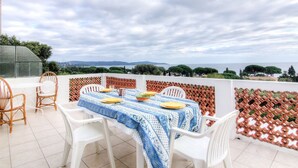 A terrace with a table set for four, surrounded by wicker chairs and white plastic chairs, overlooking a scenic landscape.