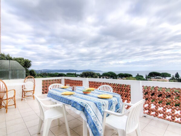 A terrace with a table set for four, surrounded by wicker chairs and white plastic chairs, overlooking a scenic landscape.