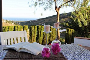Un libro abierto sobre una mesa con mantel estampado, dos candelabros con flores y una vista panorámica del paisaje costero.