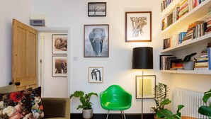 A cozy living room with a green chair, a sofa with floral cushions, a bookshelf, and various framed pictures on the wall. - Raise the Bar, by Veeve (London)