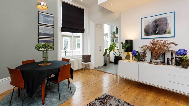 A modern dining area with a round table, chairs, and a black tablecloth. There are framed pictures on the wall, a potted plant, and a large elephant picture. - Raise the Bar, by Veeve (London)